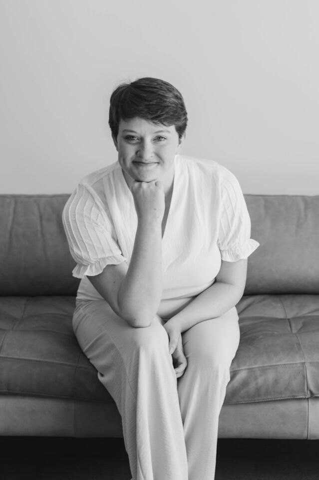 Alyssa, Newborn Care Specialist in Mesa Newborn care specialist in East Mesa seated on a sofa, smiling warmly at the camera in a relaxed, approachable pose. Light-colored outfit and minimalistic background reflect calm, professional support for families with newborns.